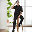 Man wearing modern black short-sleeve shirt and pants, seated indoors with city view.