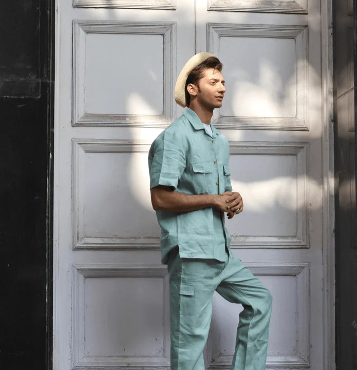 Man in mint green two-piece outfit and straw hat posing by white paneled door in sunlight