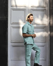Man in mint green two-piece outfit and straw hat posing by white paneled door in sunlight