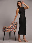 Woman in sleeveless black dress and beige heels posing beside geometric patterned chair
