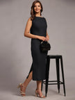 Woman in a sleeveless black midi dress with slit, holding a black purse, wearing heels