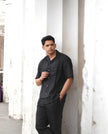 Man wearing black short sleeve shirt and trousers leaning against a white column