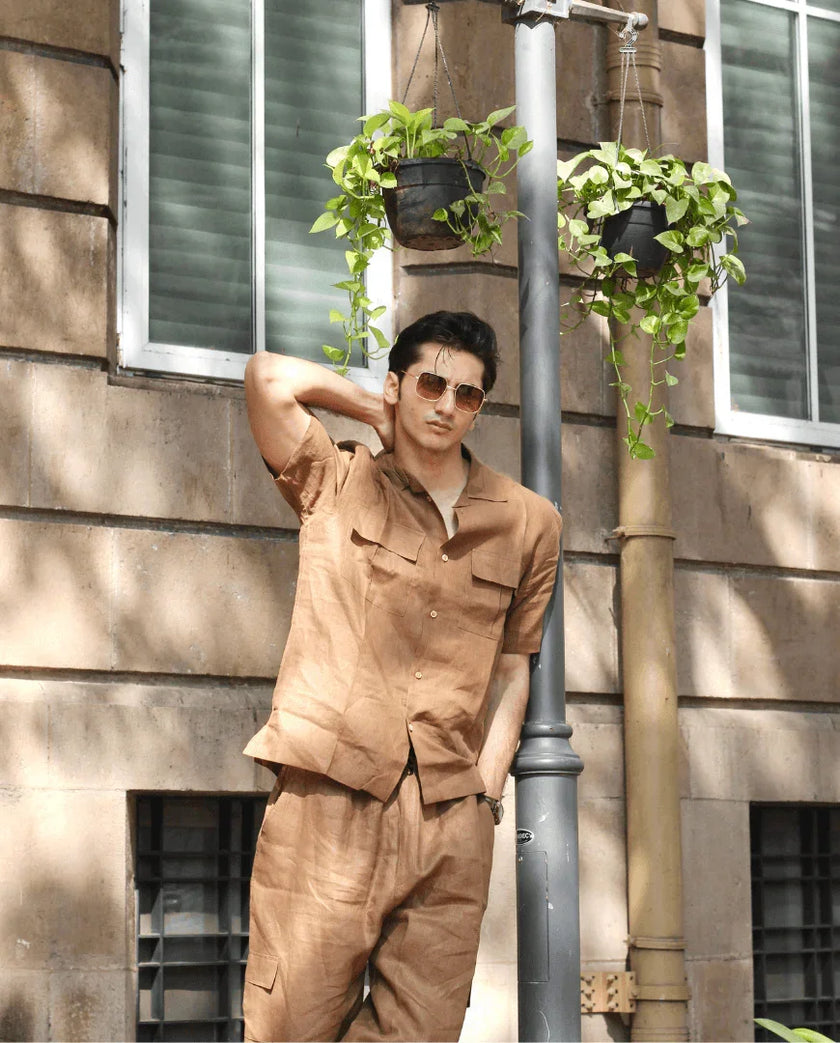 Man in brown short-sleeve shirt and trousers posing outdoors by hanging plants and windows