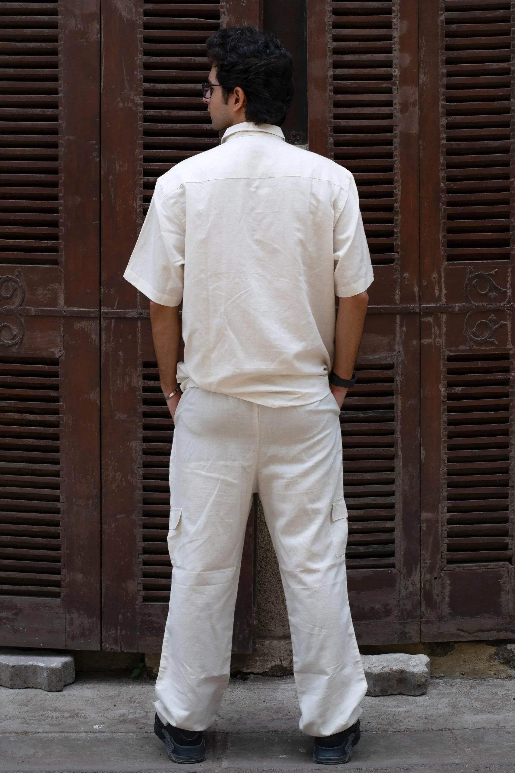Man in cream short-sleeve shirt and pants set, standing outdoors facing wooden shutters