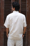 Man in cream short-sleeve shirt and pants standing with back to camera, rustic backdrop