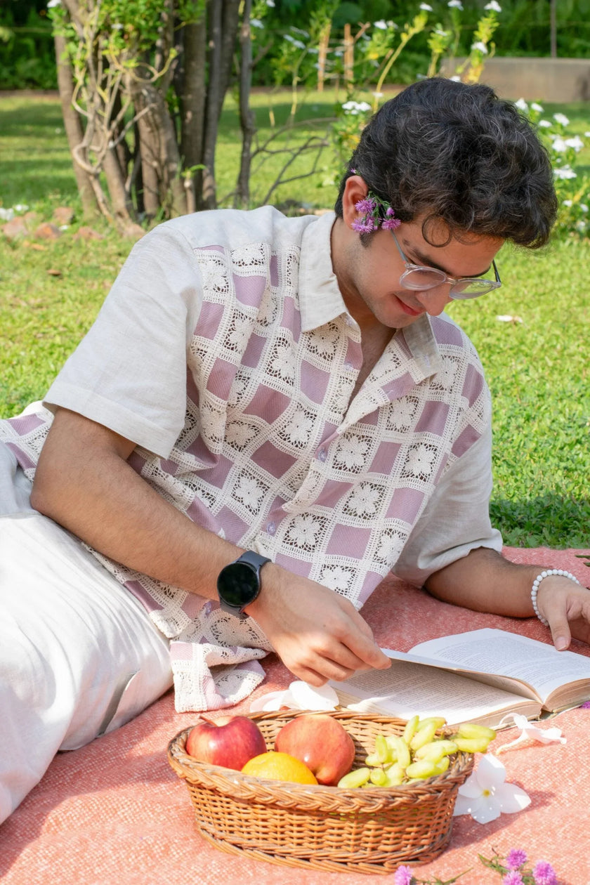 Man in crochet patchwork shirt reading a book at a picnic with fruit basket on grass