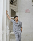 Man in a light grey short-sleeve shirt and pants set standing against a white wall
