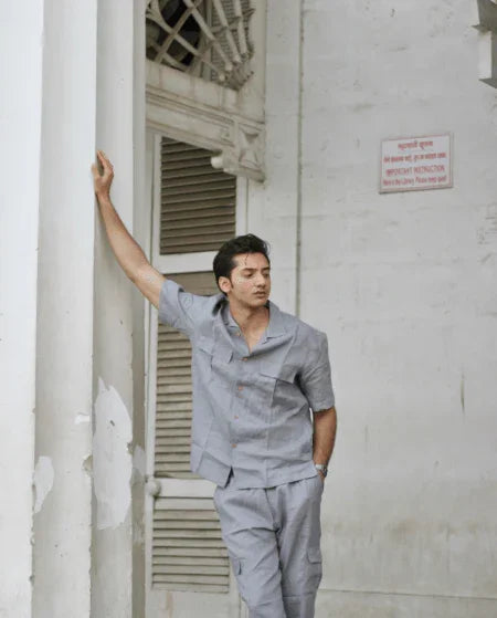 Man in a light grey short-sleeve shirt and pants set standing against a white wall