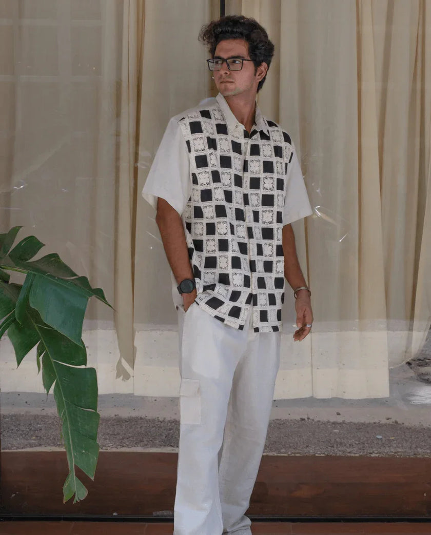 Man wearing black and white checkered short sleeve shirt and white cargo pants, standing indoors.